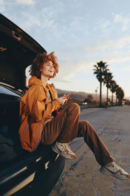 Woman lifestyle outdoors, leaning by car on city street, curly hair, warm sunset light, brown jacket and pants, relaxed pose, holding smartphone, golden hour, palm trees blur