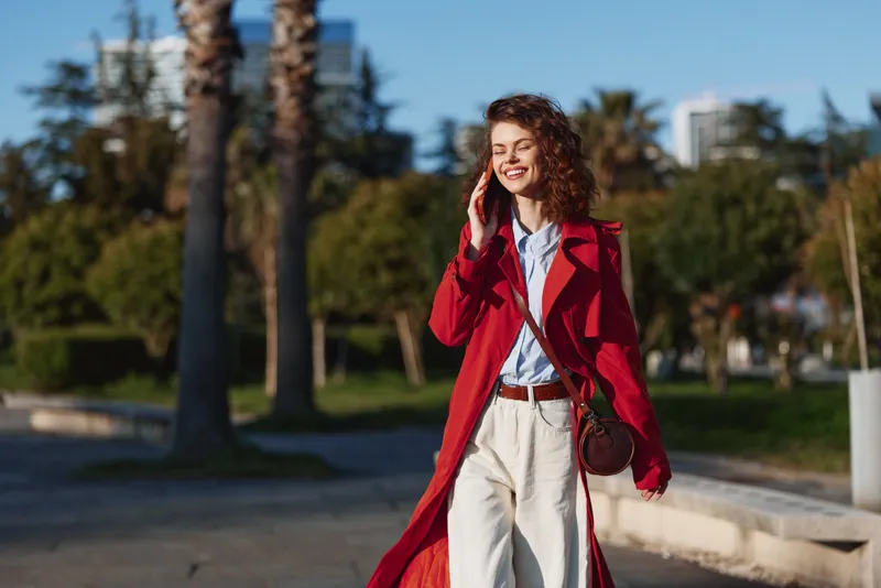One stylish young woman with curly hair walks outdoors in a bright red coat along a sunny boardwalk, smiling softly as palm trees and a modern skyline frame her, while casual attire and a warm glow