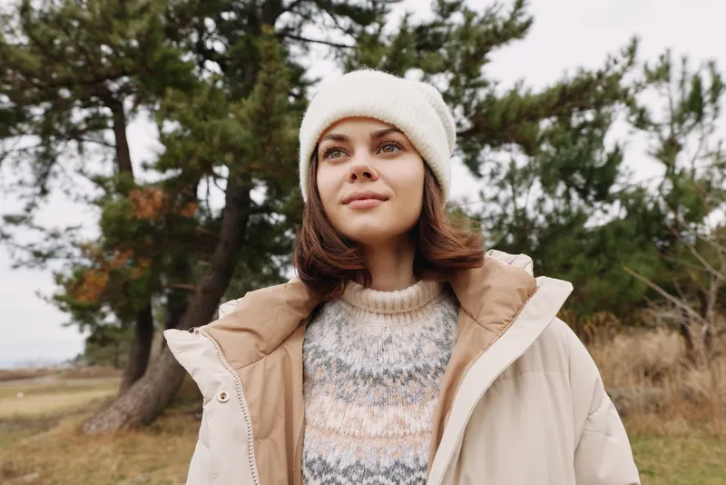 Woman in beige coat and knit sweater stands outdoors among trees, crisp weather and calm portrait highlighting contemporary fashion and seasonal style