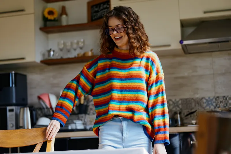 A joyful woman in a colorful striped sweater stands in a bright kitchen, smiling as she leans on a chair. Casual home scene conveying warmth, comfort, and everyday style.