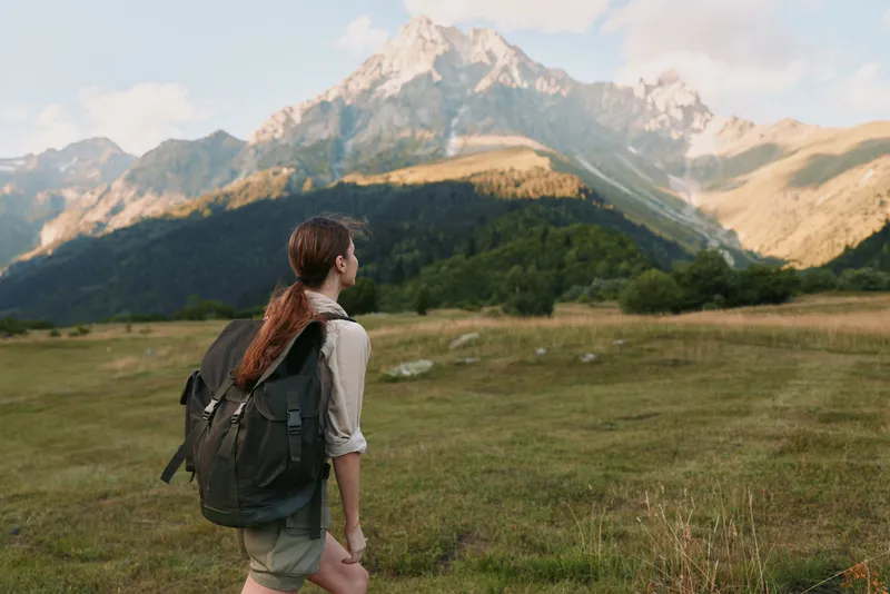 Woman hiker with backpack stands in a wide alpine meadow, gazing at distant mountains, enjoying outdoor lifestyle and nature in a scenic landscape.