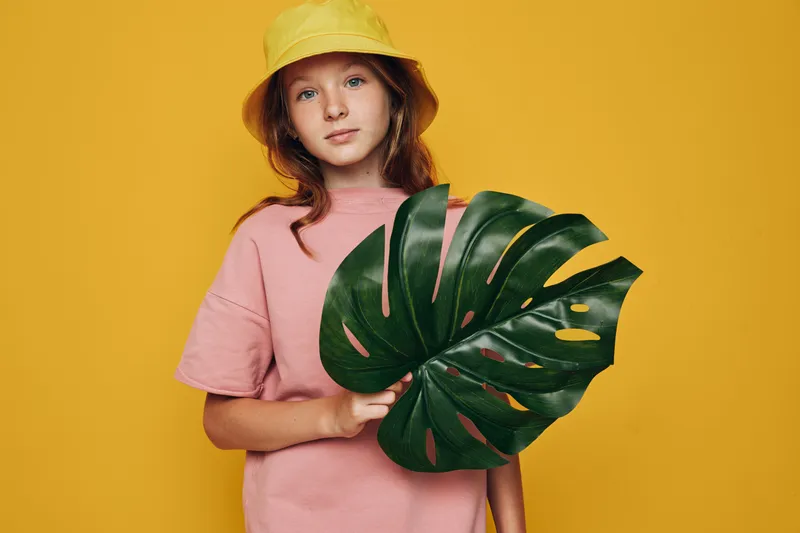 Young girl in a yellow hat holding a large green tropical leaf against a yellow background. Fashion portrait with bright colors and natural elements.