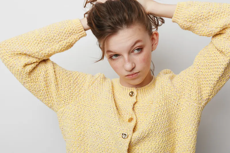 Young woman in yellow knitted sweater adjusts her hair with both hands, looking sideways. Casual style, natural light, indoor portrait with neutral background and relaxed mood.