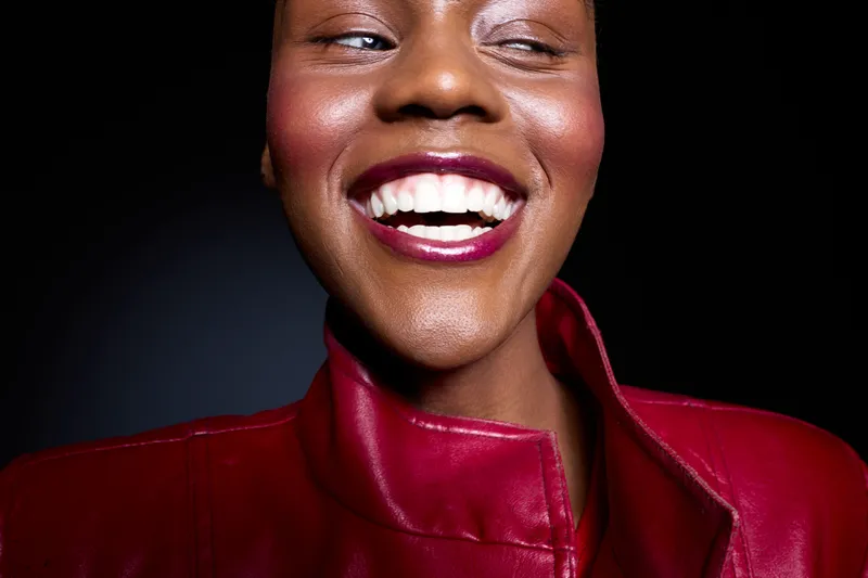 Close-up of happy dark skinned African woman smiling with white teeth wearing red leather jacket. Studio portrait with black background. Concept of joy, beauty, fashion, and confidence.