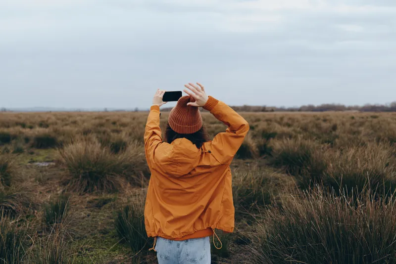 Woman lifestyle outdoors in a grass field wearing an orange jacket holding a hat overhead in autumn light Soft clouds suggest calm freedom and relaxed posing for everyday life