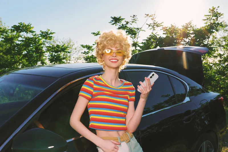 Smiling woman with curly hair wearing a striped shirt stands beside a sleek car under bright sun. Retro color palette enhances a playful lifestyle mood and carefree outdoor moment.