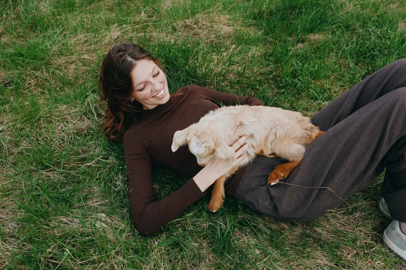 Cheerful woman outdoors, lying on grass, softly stroking a playful goat kid, warm sunlight, relaxed pose, gentle breeze, rustic charm, wholesome scene, affectionate moment, natural colors
