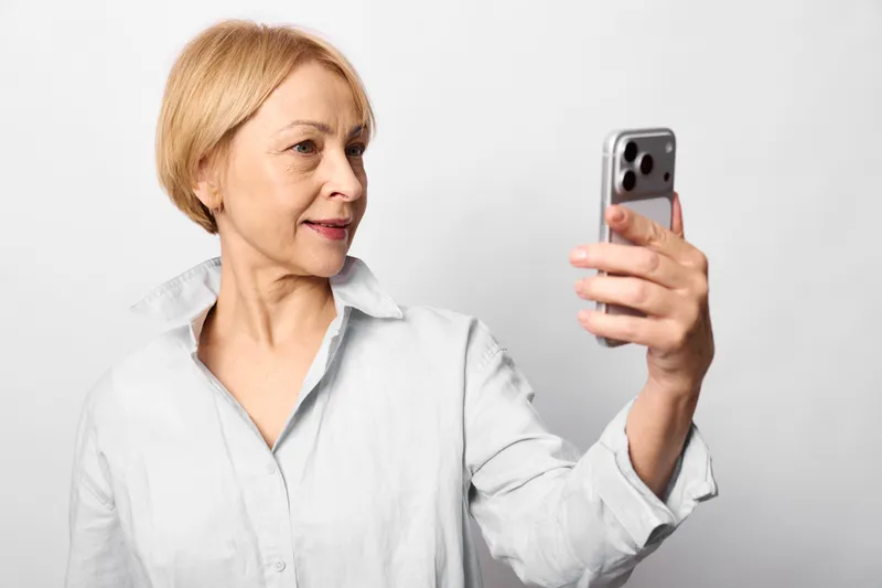 Woman elderly smartphone selfie indoors communication technology portrait, senior woman holding mobile phone, slight smile, white studio background, relaxed posture, close view