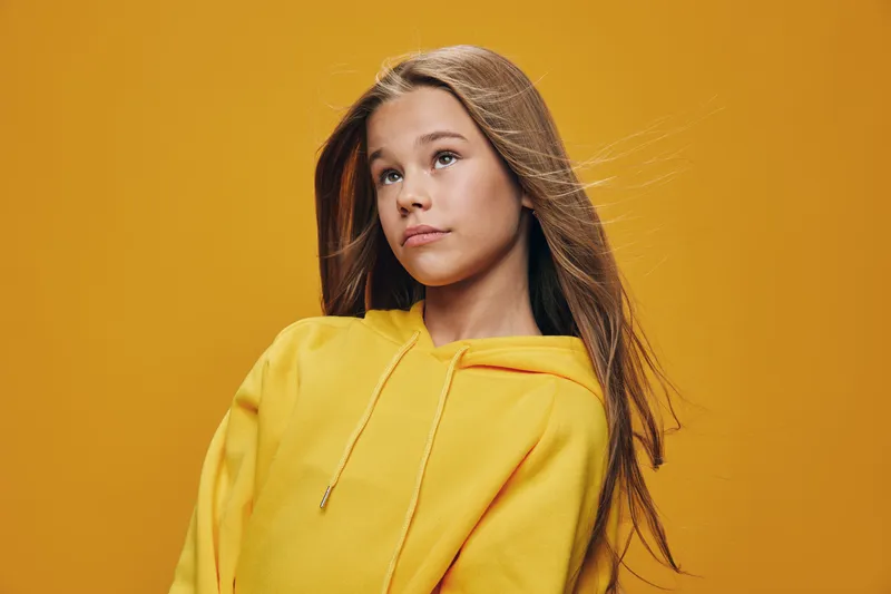 Teenage girl with long hair wearing yellow hoodie standing against yellow background looking away with serious expression. Studio portrait of young female model in casual clothes.