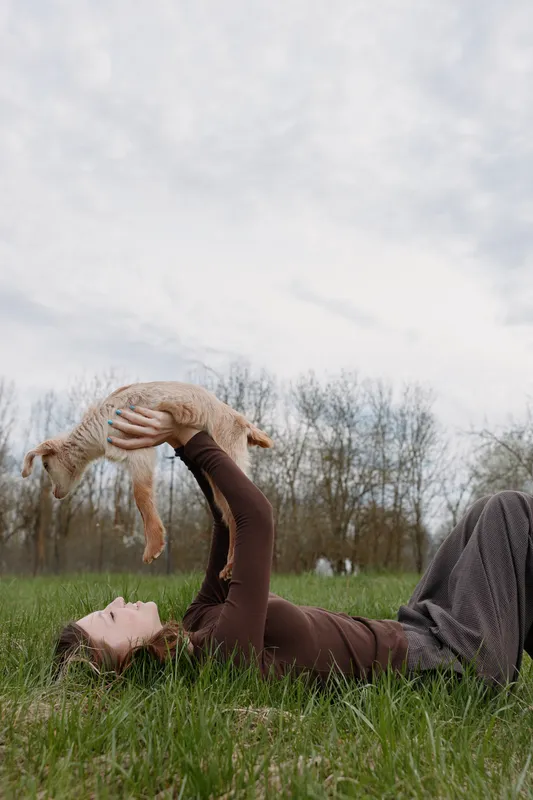 Relaxed woman lying on grass lifts a playful goat kid outdoors, capturing a warm lifestyle moment and close bond with animals in a serene countryside.