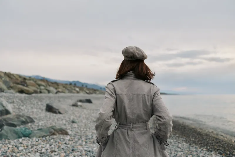 Woman wearing coat and beret stands on rocky beach looking at cloudy sea horizon. Concept of autumn fashion, solitude, and peaceful outdoor lifestyle near water.