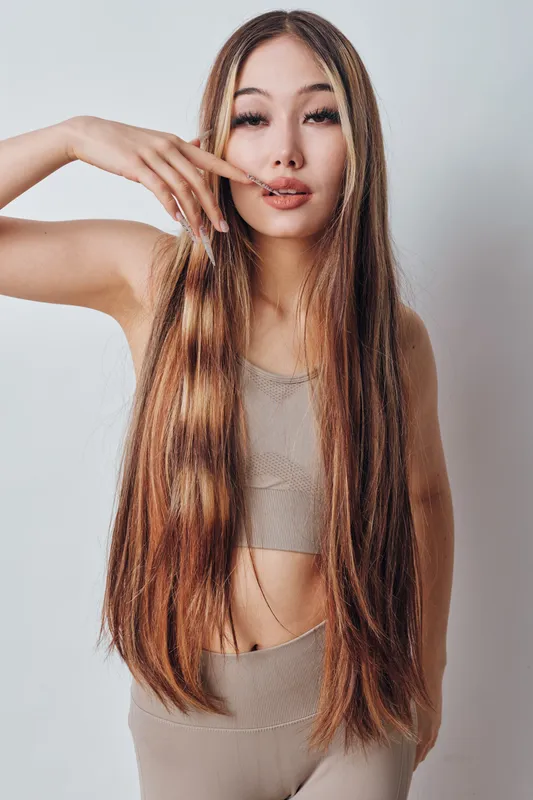 Young woman with long straight hair wearing beige top and pants posing with hand near face against white background, beauty, fashion, lifestyle, studio portrait.