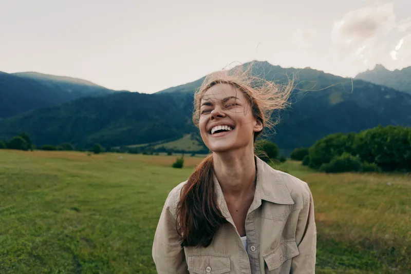 Happy woman enjoying outdoor life, smiling in a grassy field with mountains in the background, casual jacket, sunlit moment, lifestyle portrait of carefree everyday joy