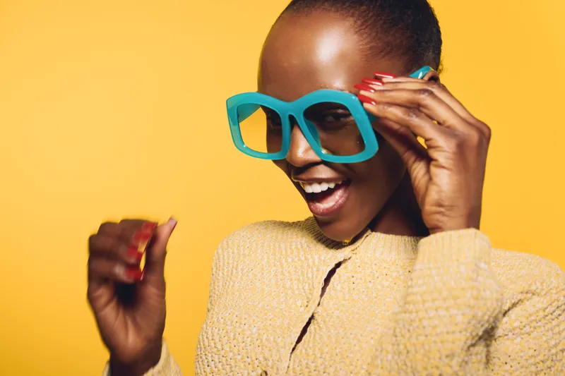 Happy dark skinned African woman in large blue glasses smiles joyfully wearing yellow sweater on yellow background. Concept of fashion, beauty, and positive mood with vibrant colors.