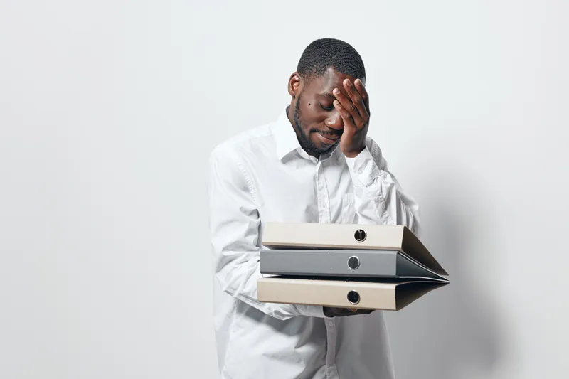 Black man with dark skin holding folders and covering face with hand, showing stress or frustration in office environment with white shirt and neutral background.