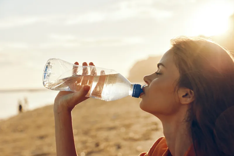 Woman drinking water from a plastic bottle outdoors in warm sunlight, summer day, athletic posture, focused face, hydration for health, clear sky background, natural colors, after workout