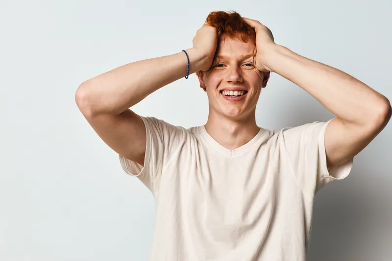 Young man with red hair smiling happily and holding his head with both hands in casual white t shirt against plain background.