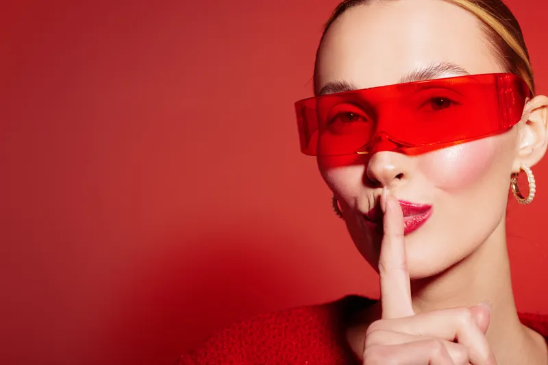 Young woman in red sunglasses and sweater makes shushing gesture with finger on lips against red background. Concept of secrecy, silence, and mystery in fashion portrait.