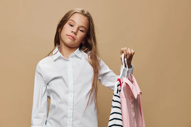 Girl wearing white shirt holding colorful clothes on hangers looking thoughtful isolated on beige background, casual fashion and clothing choice concept for young person.