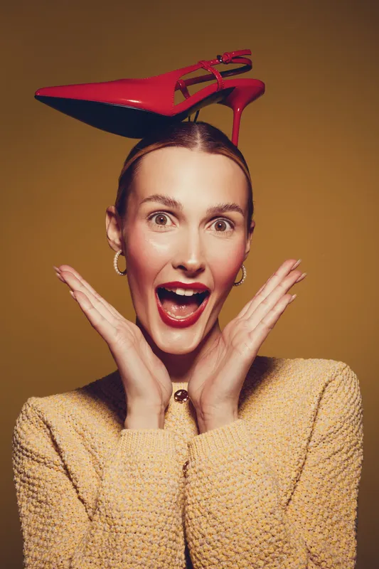 Surprised woman with red lipstick and hoop earrings holds red high heel shoe on her head. She wears a yellow sweater and poses against a brown background with expressive facial expression.
