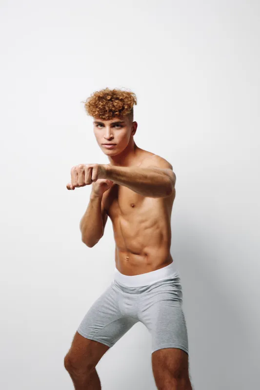 Young athletic man with curly hair practicing boxing stance shirtless in gray shorts on white background for fitness training and workout session.
