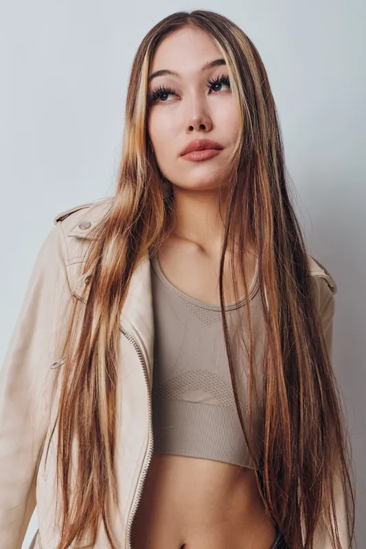 Young woman with long hair wearing beige jacket and crop top standing against white background looking away with serious expression and natural makeup.