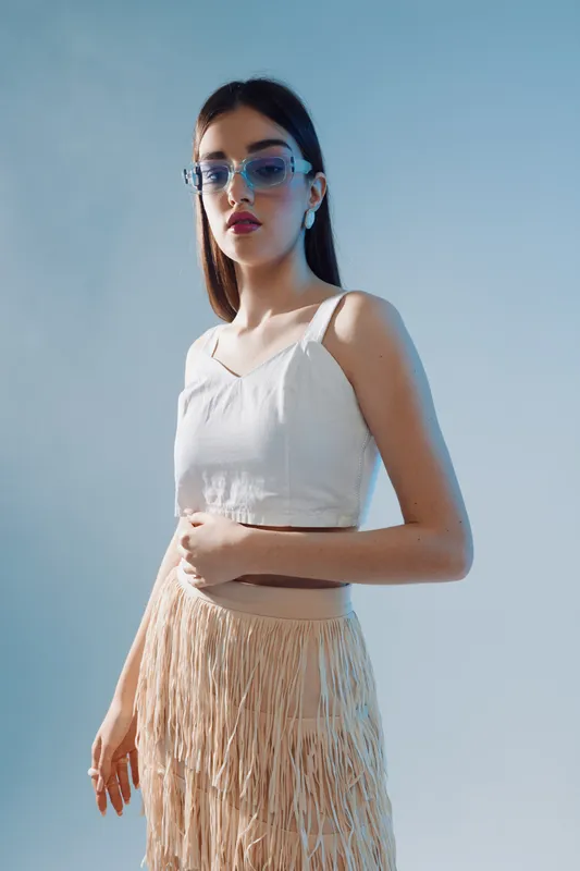 Young woman in a white top and fringed skirt poses confidently wearing blue sunglasses. Studio shot on a light blue background. Concept of woman beauty fashion and style.