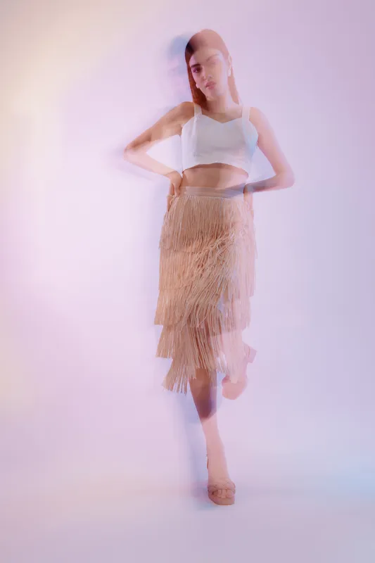 Young woman in a white crop top and fringed skirt poses with a soft motion blur effect. Studio shot on a pastel background. Concept of woman beauty fashion and movement.