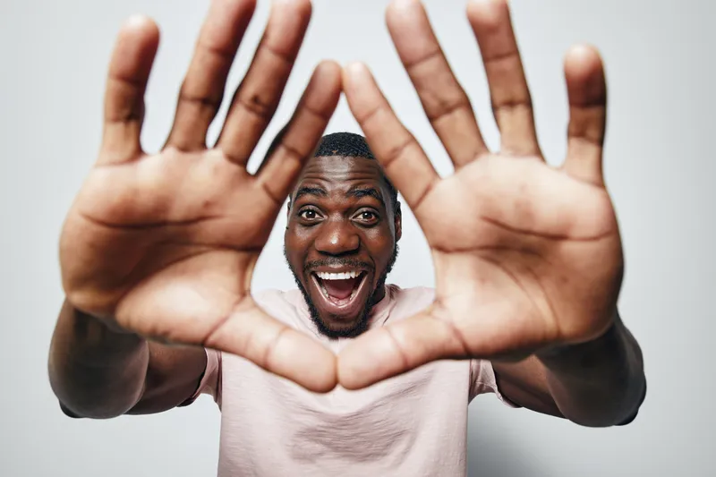 African man with dark skin and short hair smiling and showing hands close to camera in gesture on white background. Happy male portrait with open mouth and casual t shirt.