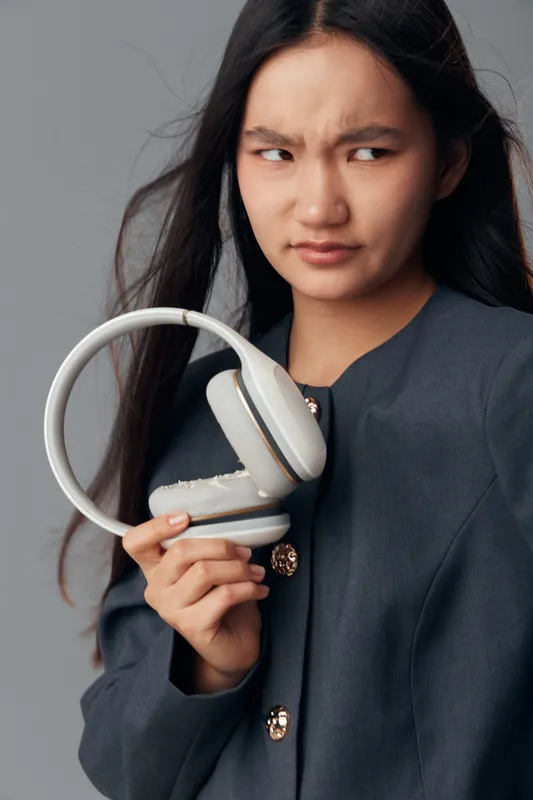 Woman holding broken wireless headphones with confused expression. Asian female in dark jacket examining damaged audio device in studio with neutral background. Technology and lifestyle concept.