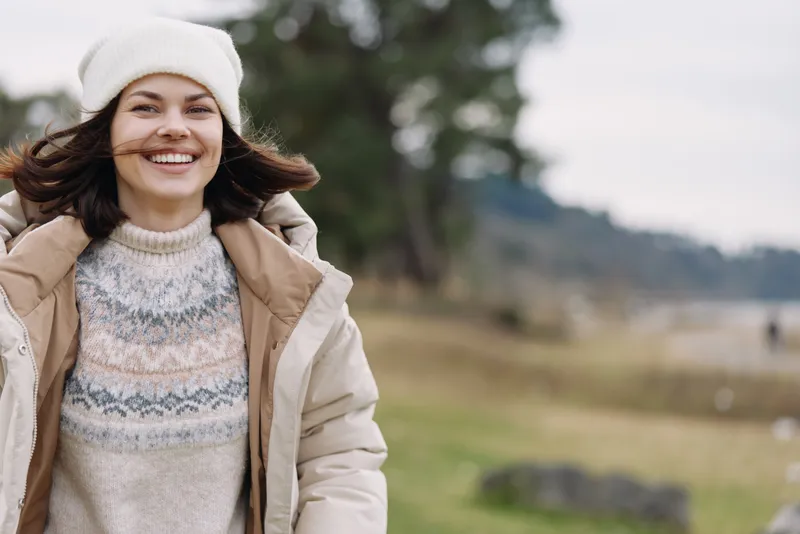 Smiling woman in a warm winter coat outdoors, natural light, breeze, countryside setting with trees and open space, casual portrait in a peaceful outdoor scene