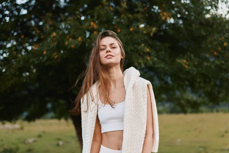 A young woman stands outdoors in a park, wearing a white crop top and a white knit wrap, exuding relaxed confidence and casual wellness in the sunlight.
