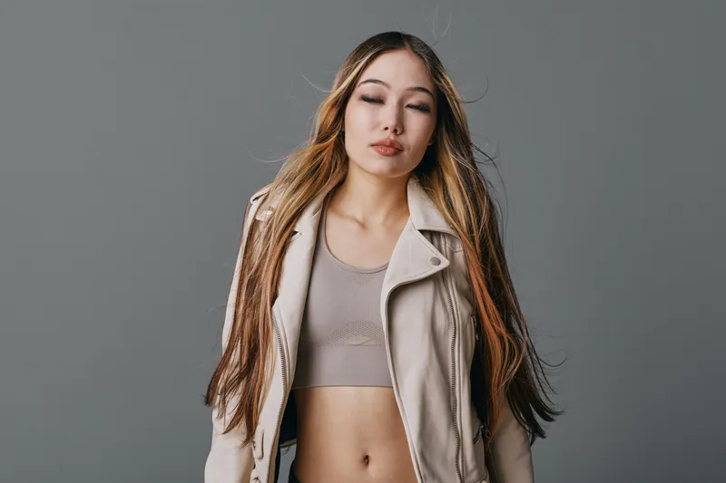 Young woman with long hair and closed eyes wearing casual jacket and crop top standing against grey background portrait.