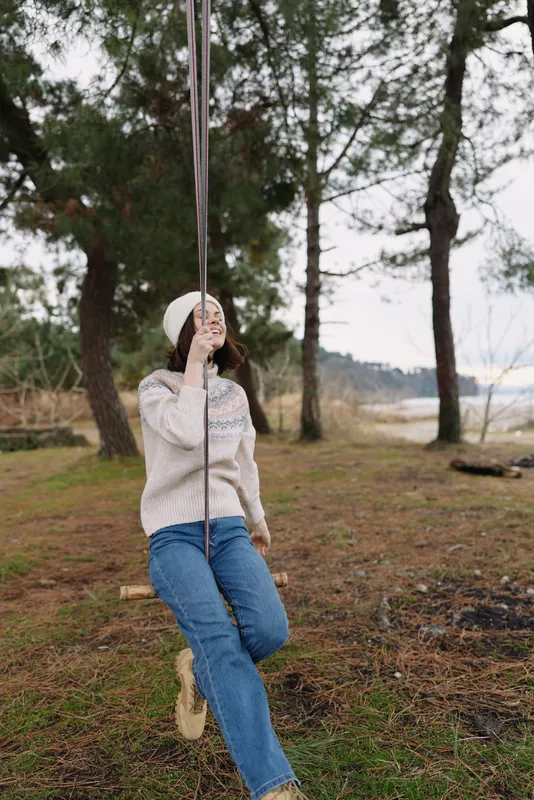 Woman on a swing in a pine forest, casual sweater and jeans, enjoying outdoor leisure, nature scenery, peaceful moment with tall trees and open field