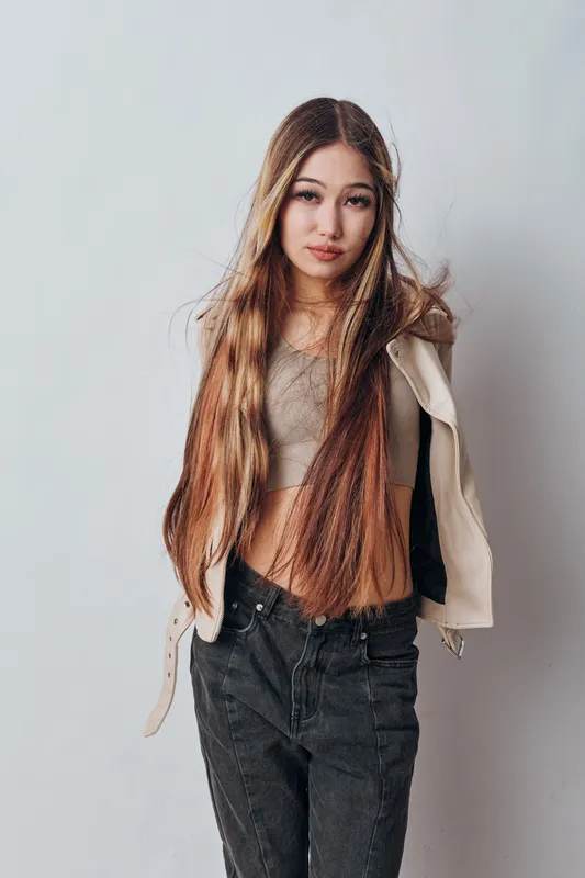 Young woman with long brown hair wearing casual beige jacket and gray top posing in front of gray background. Model standing relaxed with hands behind back for portrait photography.