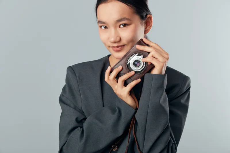 Young woman posing with vintage camera in hands wearing oversized gray blazer against neutral background for photography and fashion concept portrait.