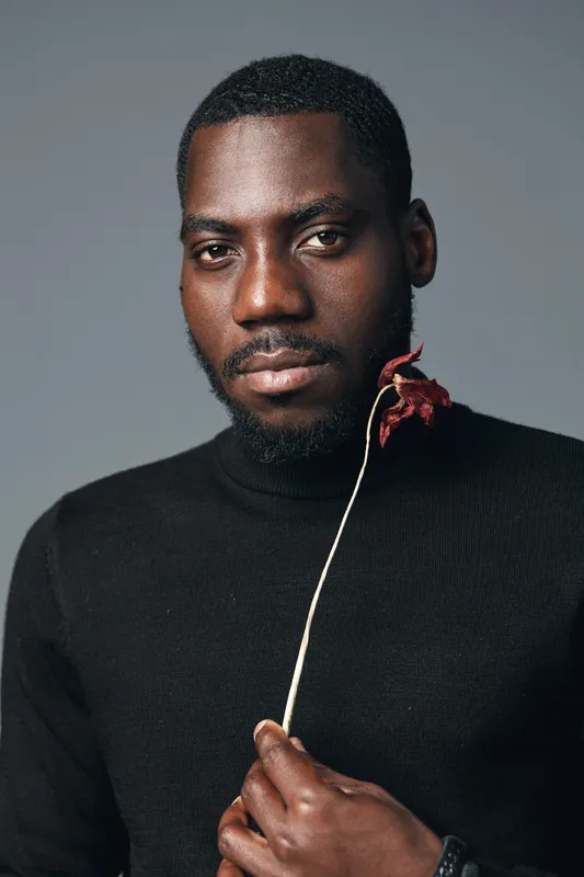 African man with dark skin wearing black turtleneck holding dried flower in creative conceptual visual style portrait with serious expression on gray background.