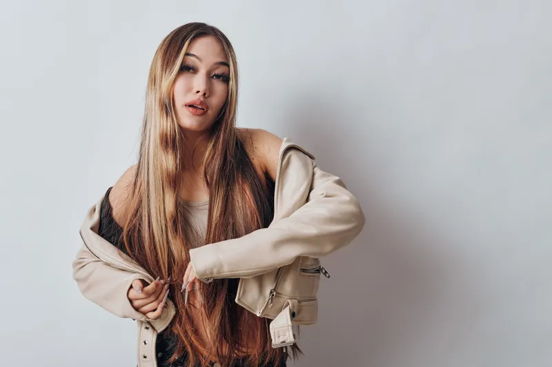 Woman with long hair posing in beige jacket over shoulders against white background, looking at camera with relaxed expression and casual style in studio setting