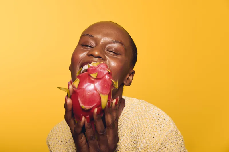Happy African woman with dark skin holding a dragon fruit close to her face, smiling with eyes closed. Bright yellow background and casual beige sweater. Concept of joy and healthy lifestyle.