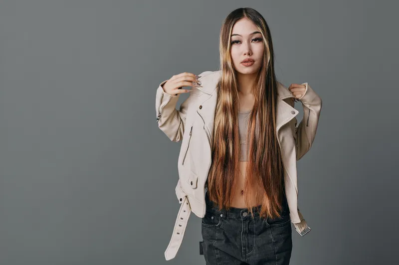 Young woman with long braided hair wearing casual jacket and crop top posing confidently against grey background in studio for fashion portrait photography.