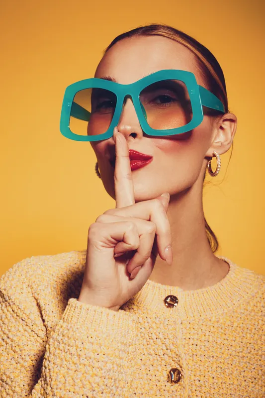 Stylish woman with oversized turquoise sunglasses and red lipstick makes shushing gesture with finger on lips. Yellow background and knitted sweater create vibrant fashion portrait.