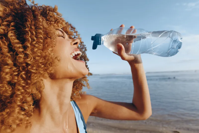Smiling woman with curly hair drinking water from a plastic bottle, outdoors near the sea, summer wellness concept, hydration and active lifestyle, bright natural light, side view