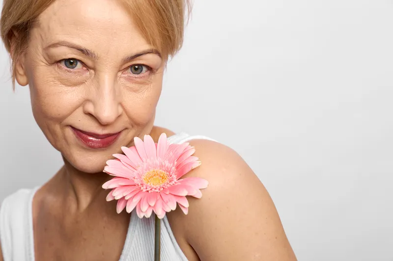 Woman portrait older smiling, holding pink gerbera flower near shoulder, gentle studio light, calm expression, soft focus, white background, closeup framing, natural skin texture