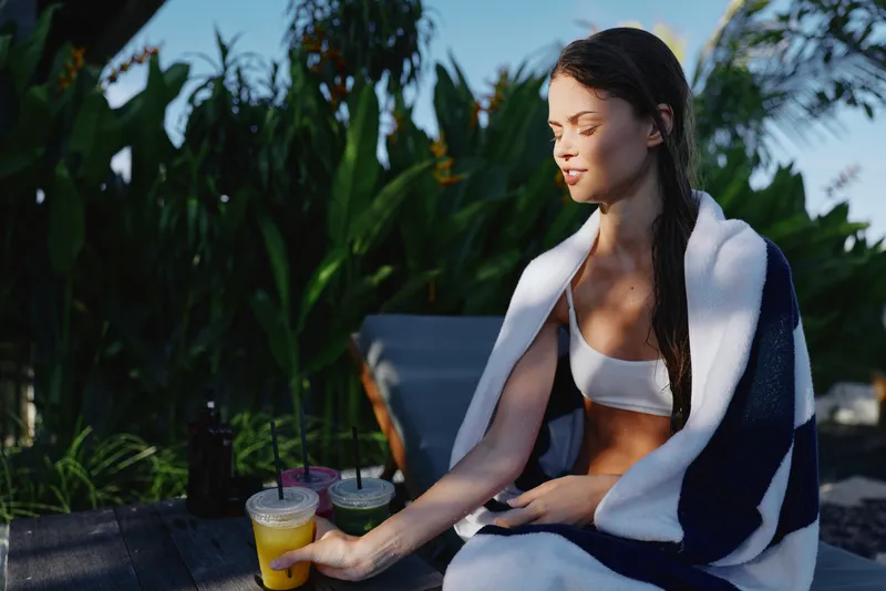 Cheerful woman outdoors, wrapped in a towel, sits beside a table with colorful beverages, sunlit moment, relaxed posture, lush greenery background, casual lifestyle mood