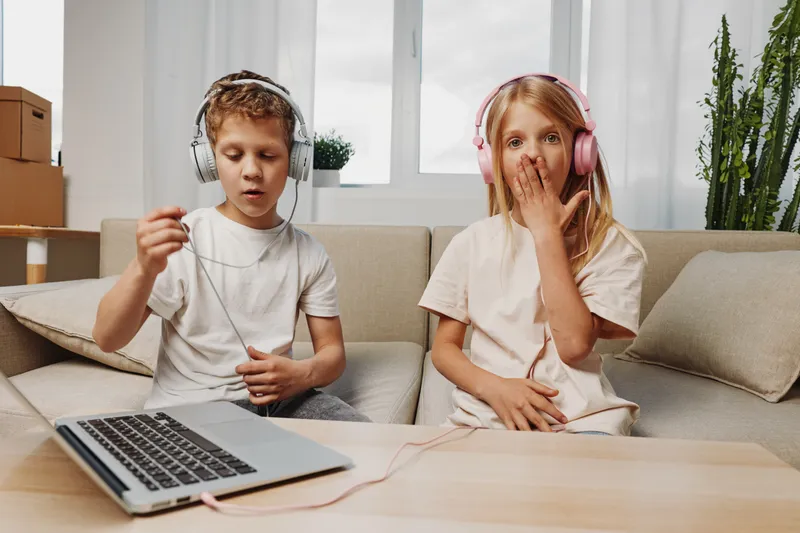 Two children wearing headphones sit on a beige sofa with a laptop on a wooden table, enjoying music or a game. Bright room with natural light and cozy home atmosphere.