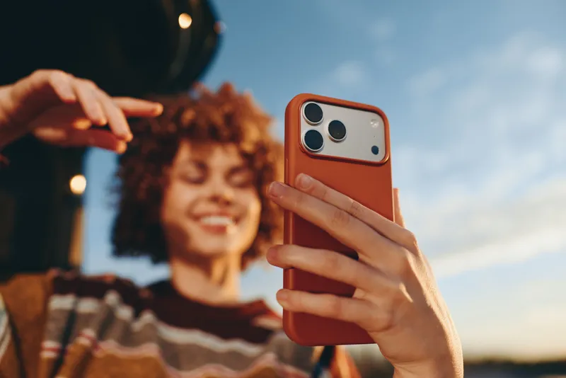 Woman lifestyle outdoors hands hold a smartphone taking a selfie, smiling friends appear blurred in background, warm sunset sky and casual leisure mood create cheerful moment today