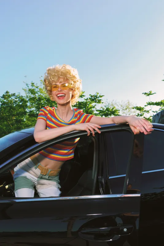 Young woman with curly blonde hair leans from a sleek black car, wearing a colorful striped top. Sunny outdoor scene captures carefree summer lifestyle and retro charm.