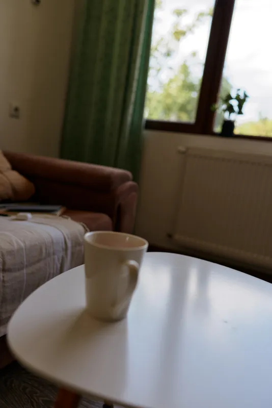 A white ceramic mug sits on a round coffee table in a cozy living room, with a sofa, cushions, and a sunlit window framing greenery outside, creating a calm home moment.