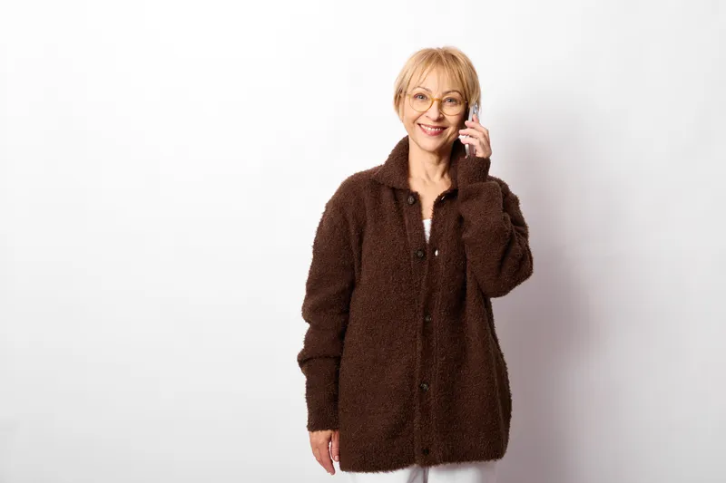 Smiling woman portrait indoors, wearing glasses and a brown fleece jacket, short blonde hair, relaxed pose, studio white background, soft light, calm mood