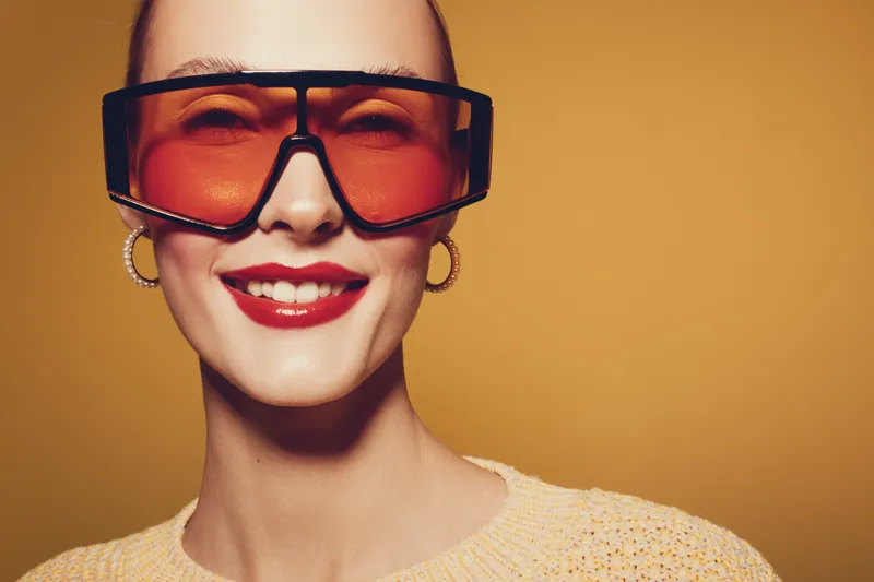Smiling woman with red lipstick wearing large orange sunglasses and hoop earrings poses against yellow background. Concept of fashion, style, confidence, and modern accessories.